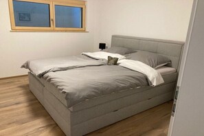 Ein Einzelbett mit grauem Kopfteil und grauer Matratze, ein Nachttisch mit Lampe sowie ein Fenster mit Jalousien.