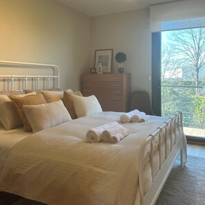 A neatly made bed with rolled towels, a dresser with a framed picture, and a view of trees through a window.