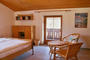 Ein Zimmer mit einem Bett, einem Korbstuhl, einem kleinen Tisch, einem Bücherregal und einer Tür, die auf einen Balkon führt.