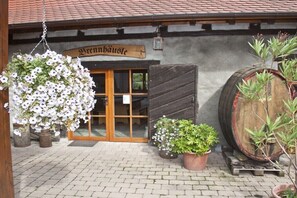 Ein traditionelles Gebäude mit einem Holzschild, auf dem 'Brennhausle' steht, einem großen Holzfass und Kübelpflanzen.