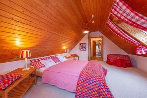 Ein gemütliches Schlafzimmer im Dachgeschoss mit schrägen Wänden, einem Bett mit pinker Decke, einem kleinen Tisch mit Lampe und einer rot karierten Tischdecke.