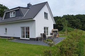 Ein zweistöckiges Haus mit geneigtem Dach, großen Fenstern und einer Terrasse mit Gartenmöbeln.