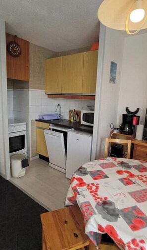 Eine kompakte Küche mit Herd, Mikrowelle und Geschirrspüler, ein Holztisch mit Blumenmuster-Tischdecke und ein Bett mit rotem und weißem Bezug.