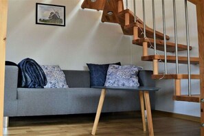 Ein Zimmer mit grauem Sofa, gemusterten Kissen, einem kleinen Tisch und einer Wendeltreppe aus Holz.