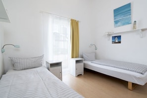 Ein Zimmer mit zwei Betten, einem Schreibtisch, einem Stuhl, einem Fenster mit Vorhängen und einem Bild an der Wand.
