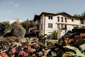 Ein zweistöckiges Gebäude mit einem Balkon, umgeben von üppigem Grün und blühenden Pflanzen.