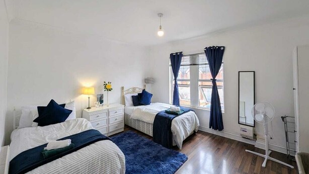 A bedroom with two beds, a bedside table with a lamp, a mirror, a standing fan, and a window with curtains. - Contractor Business Family Stay Near Southampton Airport 1BR (Eastleigh)