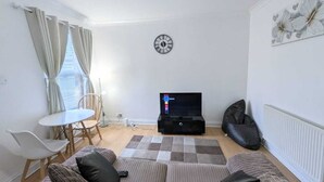 A living room with a sofa, a coffee table, a TV, a clock, and a radiator. - Contractor Business Family Stay Near Southampton Airport 1BR (Eastleigh)