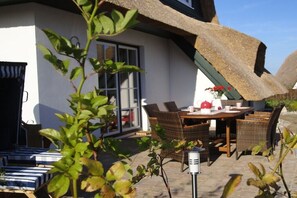Eine Terrasse mit Tisch und Stühlen, einem Reetdach und einem weißen Gebäude mit Fenstern.