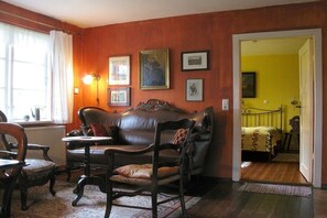 Ein Zimmer mit einem braunen Ledersofa, Holzstühlen, einem kleinen Tisch und einem Heizkörper. An der Wand befinden sich gerahmte Bilder und eine gelbe Tür, die in einen anderen Raum führt.