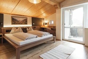 A bedroom with a wooden ceiling, a large bed with pillows, a nightstand, a wall-mounted lamp, and a view through a glass door. - Holiday apartment with 1 bedroom (Telfes im Stubaital)