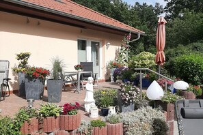 Ein Haus mit einem Ziegeldach, einer Terrasse mit Topfpflanzen, einer Statue und Gartenmöbeln.