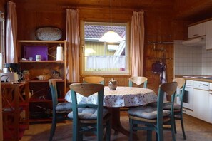 Ein Holzinterieur mit einem Esstisch, Stühlen, einem Fenster mit Vorhängen, einer Küchenzeile und einem Regal mit verschiedenen Gegenständen.