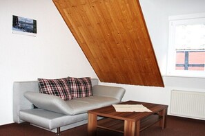A room with a slanted wooden ceiling, a grey sofa with plaid cushions, and a wooden table. - Dr. Schuldt, Hans-Otto (Waren)