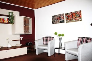 A modern living room with a white cabinet, two grey armchairs, a small table with a vase, and two wall-mounted paintings. - Dr. Schuldt, Hans-Otto (Waren)