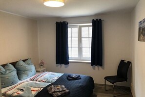Ein Schlafzimmer mit einem Bett, einem Sessel, einem Fenster mit Vorhängen und einem Nachttisch mit Lampe.