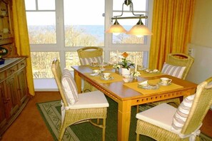 Ein Essbereich mit Korbstühlen, einem Holztisch und Blick ins Freie durch große Fenster.