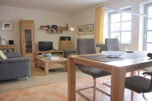 Ein Wohnzimmer mit einer Couch, einem Fernseher, einem Esstisch mit Stühlen und einem Fenster mit Vorhängen.