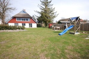 Ein Wohngrundstück mit einem rot-weiß gestrichenen Haus, einer hölzernen Spielanlage mit Rutsche und einem großen Baum.