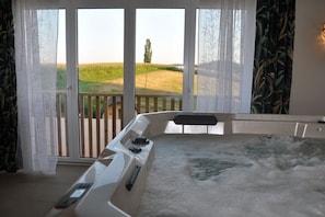 A hot tub with a control panel, a view of a grassy field through large glass doors, and a wooden deck outside.