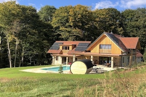 A two-story wooden house with a swimming pool, solar panels, and a barrel in the foreground.