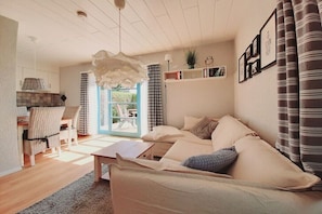 Ein Wohnzimmer mit einer Couch, einem Couchtisch, einem Essbereich mit Stühlen, einer Küchenzeile mit Herd und Blick durch eine Glastür.