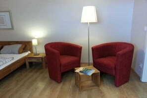 Ein Zimmer mit einem Bett, zwei roten Sesseln, einem kleinen Holztisch mit einer Zeitschrift, einer Stehlampe und einem Nachttisch mit einer Lampe.