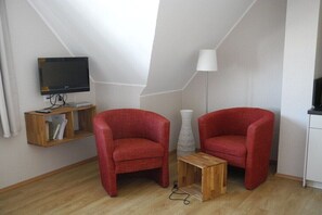 Ein Zimmer mit Fernseher auf einem Regal, zwei rote Stühle, einem kleinen Holztisch und einer Stehlampe.