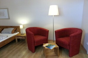 Ein Zimmer mit einem Bett, zwei roten Sesseln, einem kleinen Holztisch mit einer Zeitschrift, einer Stehlampe und einem Nachttisch mit einer Lampe.