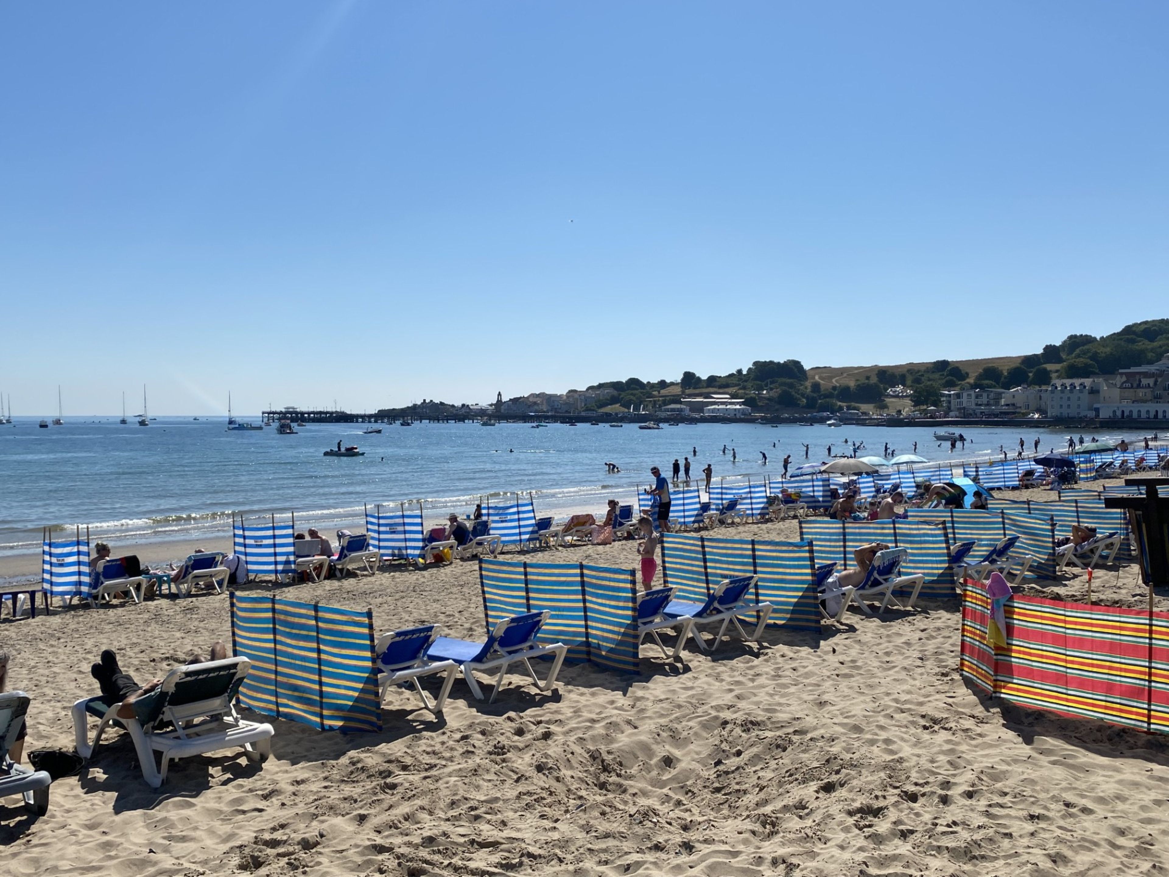 Ein Strand mit mehreren Reihen gestreifter Liegestühle und Sonnenschirme, einem klaren Himmel und Booten im Wasser.