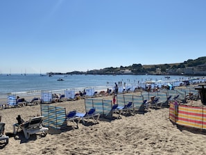 Ein Strand mit mehreren Reihen gestreifter Liegestühle und Sonnenschirme, einem klaren Himmel und Booten im Wasser.