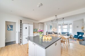 Eine geräumige Küche mit einer zentralen Kochinsel, ein Essbereich mit Tisch und Stühlen sowie ein Wohnzimmer mit blauem Sofa und Kamin.