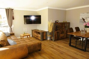 Ein Wohnzimmer mit einer Couch, einem an der Wand befestigten Fernseher, einem Holzschrank und einem Esstisch mit Stühlen.