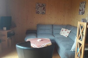 Ein Wohnzimmer mit einer Couch, einem Tisch mit kariertem Tischläufer und einem Fernseher auf einem Schrank.