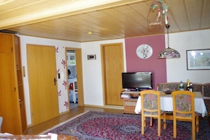 A living room with a television, a dining table with chairs, a patterned rug, wooden doors, and a ceiling fan. - Holiday apartment with 1 bedroom (Neustrelitz O.T. Klein Trebbow)