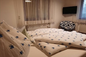 Ein Schlafzimmer mit Bett, Stuhl, Fernseher und einem fensterbeprägten Fenster mit Vorhängen.
