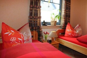 Ein Zimmer mit zwei Betten, einem Fenster mit Vorhängen, einer Pflanze auf dem Fensterbrett, einer Lampe und einer Vase mit Blumen.