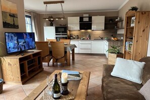 Ein modernes Wohnzimmer mit Fernseher, Holzmöbeln, einer Essecke und einer Küche mit weißen Schränken.