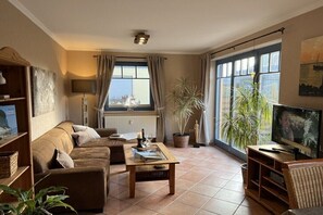 Ein Wohnzimmer mit einer Couch, einem Couchtisch, einem Fernseher und großen Fenstern mit Vorhängen.