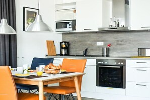 Eine moderne Küche mit weißen Schränken, einem Bosch-Ofen, einer Kaffeemaschine und einem Essbereich mit orangefarbenen Stühlen und einem Holztisch.