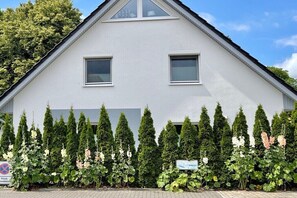 A two-story building with a triangular roof, two windows, and a row of coniferous trees in front. - Holiday home in Dahme with swimming pool (Dahme)