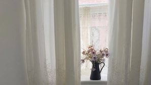 Una habitación con una cama, un ventanal con cortinas traslúcidas y un florero con flores en el alféizar.