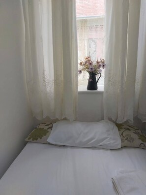 Quarto com uma cama, janela com cortinas leves e um vaso de flores no parapeito.