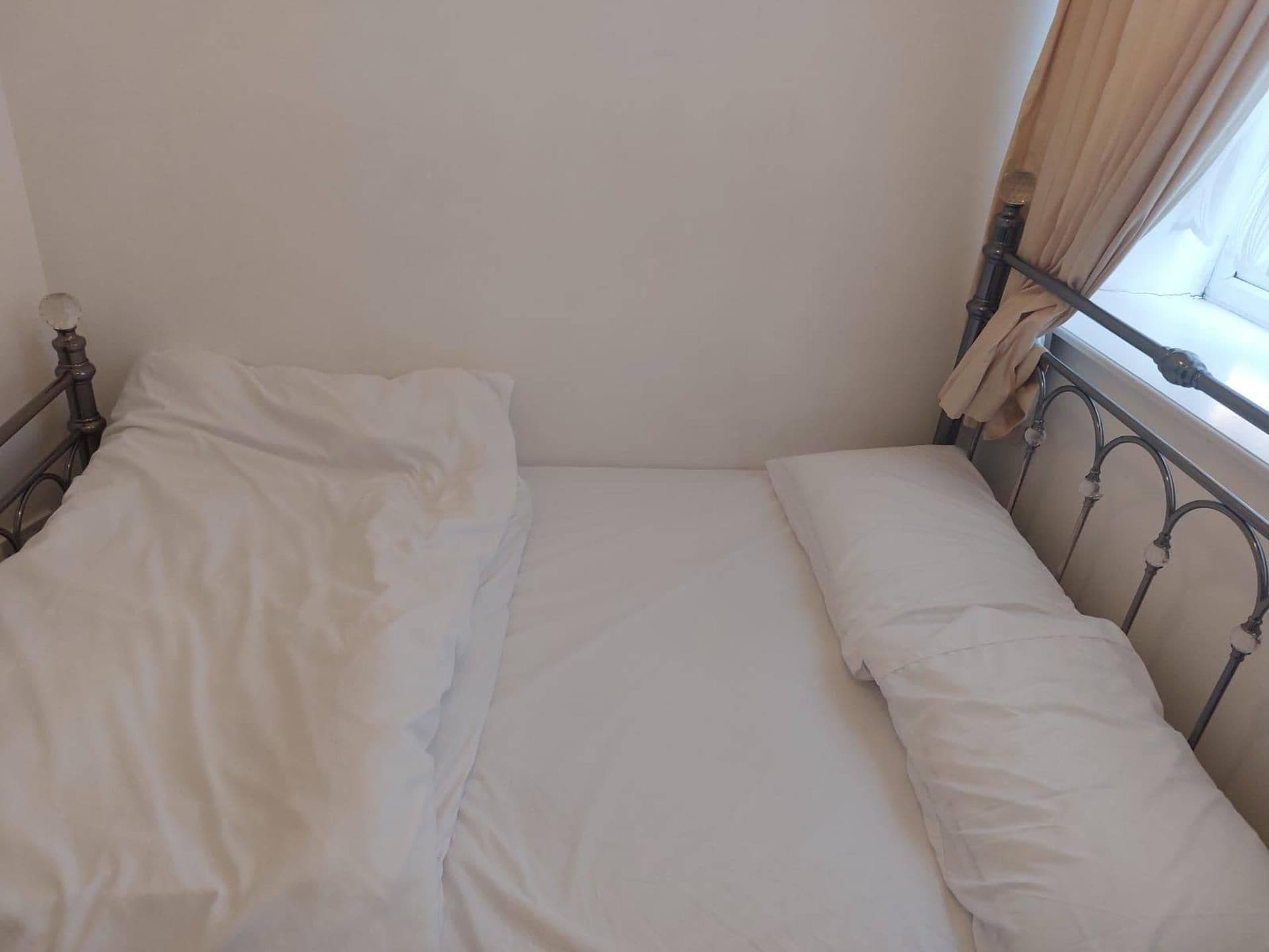 A single bed with white linens in a room with a plain wall and a window with curtains.