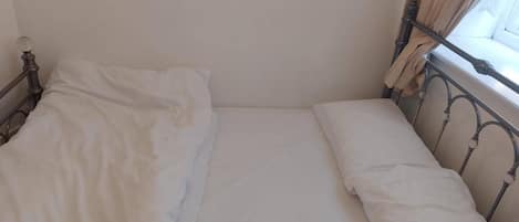 A single bed with white linens in a room with a plain wall and a window with curtains.