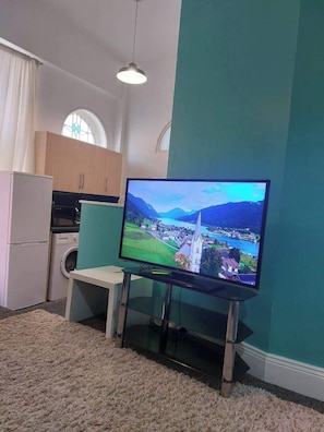 Ein Wohnzimmer mit Flachbildfernseher, Waschmaschine, kleinem Tisch und Teppichboden.