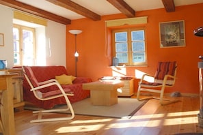 Ein Zimmer mit Holzboden, einem roten Sofa, einem Schaukelstuhl aus Holz, einem Holztisch und einem Fenster mit Jalousien.