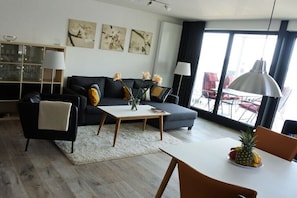 A modern living room with a sofa, coffee table, and dining table. There are abstract paintings on the wall and a large window with curtains. - Apartment in Plau am See with swimming pool (Plau am See)