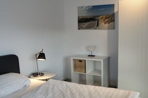 A hotel room with a bed, a bedside lamp, a small white nightstand, a wicker basket, and a framed picture on the wall. - Apartment in Plau am See with swimming pool (Plau am See)