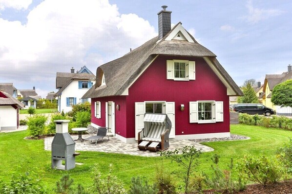 Ein rotes Haus mit einem Reetdach, einem Schornstein und einer kleinen Terrasse mit Grill und Gartenmöbeln.